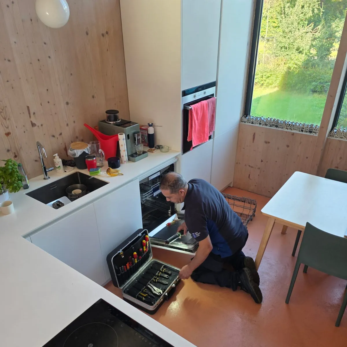 De monteur geknield voor een geopende inbouwvaatwasser in een keuken, met zijn gereedschapskoffer ernaast